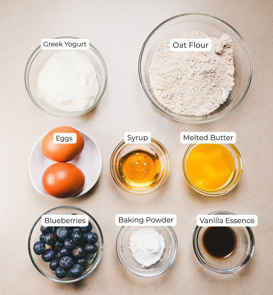 Ingredients for homemade high protein blueberry pancakes, including Greek yogurt, oat flour, eggs, blueberries, maple syrup, vanilla essence, and baking powder.
